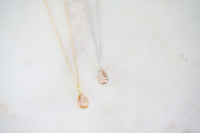 May include: Two delicate necklaces with teardrop-shaped pendants. One necklace has a gold chain and pendant, while the other features a silver chain and pendant. The pendants are a light peach color, with a textured appearance.