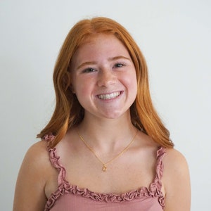 May include: A smiling person with long, red hair and a gold necklace with a small pendant. They are wearing a pink top with ruffled straps. The background is a plain white wall.
