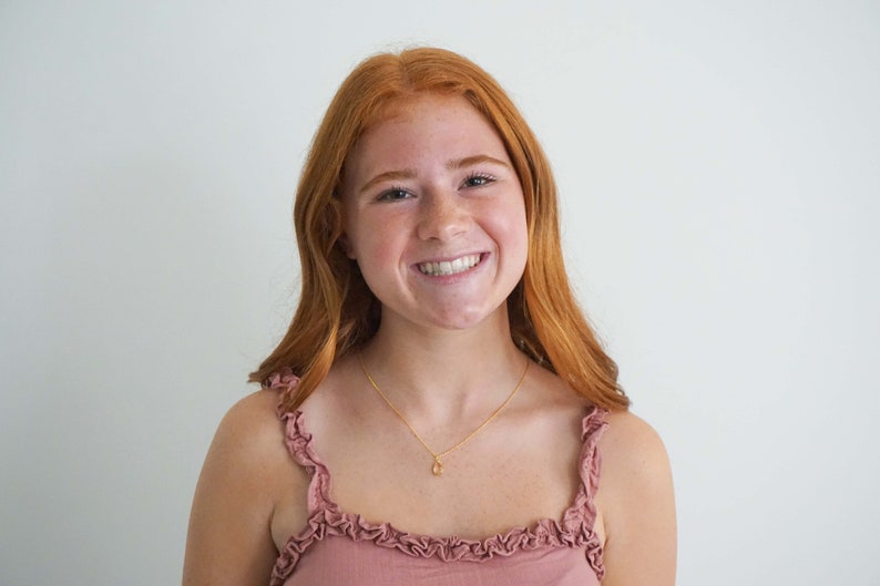 May include: A smiling person with long, red hair and a gold necklace with a small pendant. They are wearing a pink top with ruffled straps. The background is a plain white wall.