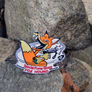 May include: Embroidered patch depicting a cartoon fox with a machine gun. The fox is orange and white, with a black outline. The patch reads "Special Force Group FOX HOUND" in red and white lettering.