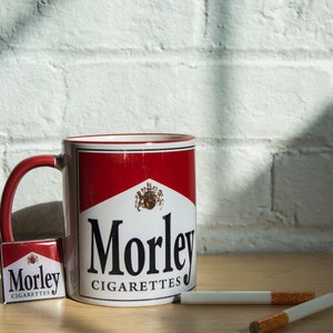 Può includere: Una tazza in ceramica bianca con un logo Morley Cigarettes rosso e bianco. La tazza ha un manico rosso ed è appoggiata su un tavolo di legno. Ci sono una piccola confezione di sigarette Morley e due sigarette sul tavolo.