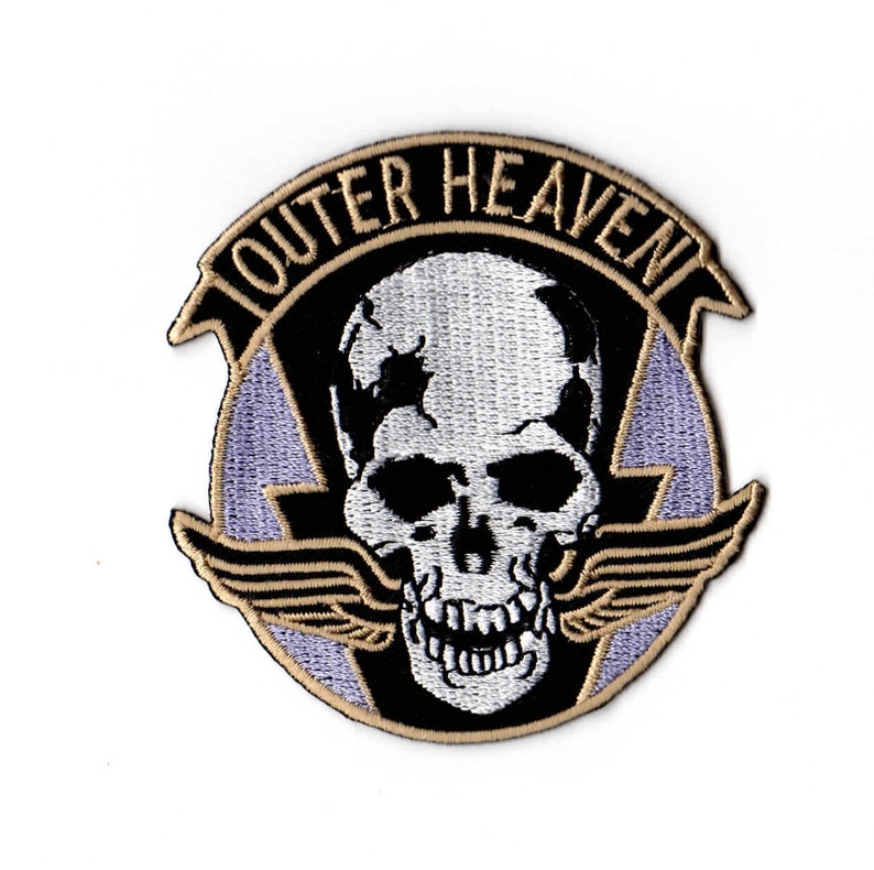 Peut inclure: Un &eacute;cusson brod&eacute; noir et blanc avec un cr&acirc;ne et des ailes. Le texte "Outer Heaven" est &eacute;crit en brun autour du cr&acirc;ne.