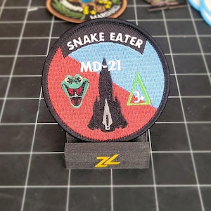 May include: A round, black-bordered patch with red, blue, and green embroidery. The patch features a black silhouette of a plane, a green snake head, and a green triangle with the number 3 inside. The text "SNAKE EATER" and "MD-21" are embroidered in white.