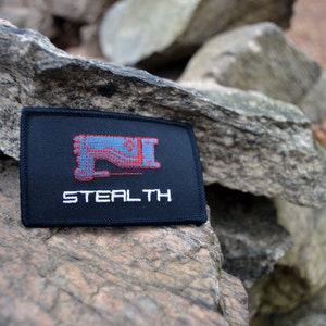 May include: Black embroidered patch featuring a red and grey graphic of a stealth aircraft and the word "STEALTH" in white text.