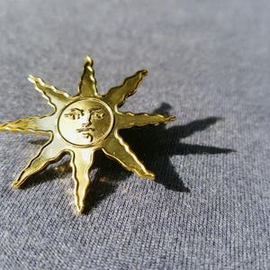 May include: A gold-colored sunburst pin with a detailed face in the center. The pin has multiple pointed rays extending outwards. The pin is set against a gray background, with the sun's shadow visible.