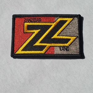 Könnte beinhalten: Ein gestickter Aufnäher in Schwarz, Gelb und Rot mit dem Text "Zanzibar" und "Land". Der Aufnäher zeigt ein stilisiertes "Z"-Design.