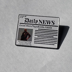 May include: A white and black enamel pin featuring a vintage newspaper headline that reads "Daily NEWS" with the subheading "Local Man Played Like Fiddle". The pin includes a black and white photo of a man with the caption "Kazuhira "Kaz" Miller, Military Strategist & Privatization Advocate".