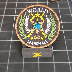 Può includere: Una toppa rotonda ricamata di colore marrone con le parole "WORLD MARSHAL" in lettere bianche. La toppa presenta uno sfondo nero con un disegno bianco e oro di un globo circondato da stelle e una corona.