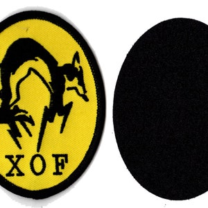 Pode incluir: Um remendo circular amarelo com uma silhueta preta de uma raposa pulando sobre um raio. O texto "XO F" está impresso abaixo da raposa.