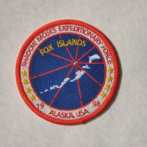 May include: An embroidered patch in red, white and blue with the text "Shadcon Moses Expeditionary Force" and "Fox Islands" in yellow. The patch also includes a map of the Fox Islands, a star design, and the text "Alaska, USA 1998".