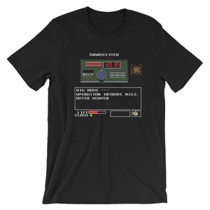 May include: Black t-shirt with a pixelated image of a video game screen. The screen displays the text "TRANSCEIVER", "BIG BOSS", "OPERATION INTRUDE N313", "OUTER HEAVEN", "LIFE CLASS", and a character's portrait.