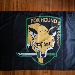 Puede incluir: Bandera negra con un emblema verde, amarillo y blanco. El emblema presenta una cabeza de zorro con un cuchillo y el texto "FOX HOUND SPECIAL FORCE GROUP".