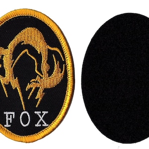 Könnte beinhalten: Ein gestickter Aufnäher in Schwarz und Gold mit einem stilisierten Fuchsdesign und dem Wort "FOX" in weißen Buchstaben. Der Aufnäher hat eine Klettverschluss-Rückseite zum Anbringen an Kleidung oder anderen Gegenständen.