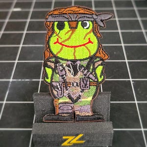 May include: A green frog wearing a brown bandana and camouflage clothing. The frog is holding a gun and has a smiling face. The frog is standing on a black stand with the letters "ZL" in yellow.