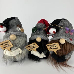 May include: Three whimsical gnome figurines with gray knit hats, each holding a wooden sign that reads "WISHING I WAS FISHING." The gnomes have different colored beards, buttons, and fishing bobber accents. One gnome has a butterfly.