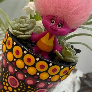 May include: A hand-painted black ceramic pot with a colorful dot design, featuring a pink Troll doll and small succulents. The pot's design includes yellow, orange, and pink circles. A whimsical decorative item.