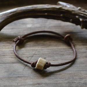 Puede incluir: Un brazalete de cuero marrón con una sola cuenta de hueso marrón claro. El brazalete está atado con un nudo a cada lado de la cuenta.