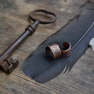 May include: Two hammered copper rings on a black feather with a vintage key and a clear quartz crystal on a wooden surface.