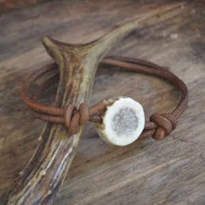 Puede incluir: Un brazalete de cuero marrón con un dije de asta de ciervo blanco. El brazalete está atado con un nudo y está sobre una superficie de madera.