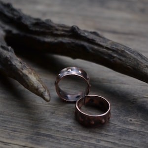 May include: Two copper rings with a hammered texture and a row of small dots around the edge. The rings are on a wooden surface.