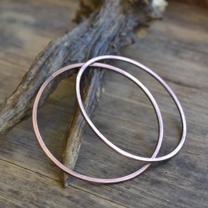 May include: Two large, round, copper hoop earrings. The hoops are thick and have a hammered texture. They are lying on a wooden surface.