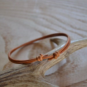 Peut inclure: Un bracelet simple en cordon de cuir brun avec deux nœuds. Le bracelet est posé sur une pièce de bois de cerf.