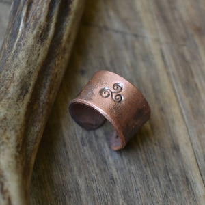 May include: A copper ring featuring a Celtic knot design. The ring is displayed on a wooden surface.