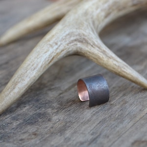 May include: A black metal ring with a copper band, resting on a wooden surface beside a white antler.