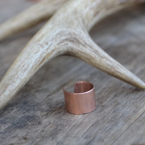 May include: A copper ring with a smooth, polished finish. The ring is lying on a wooden surface next to a white antler.