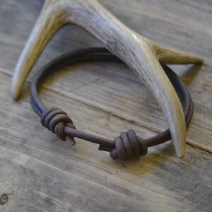 May include: A brown leather bracelet with two knots. The bracelet is lying on a wooden surface next to a white antler.