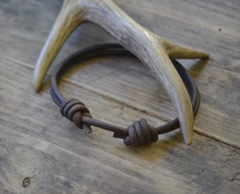 May include: A brown leather bracelet with two knots. The bracelet is lying on a wooden surface next to a white antler.