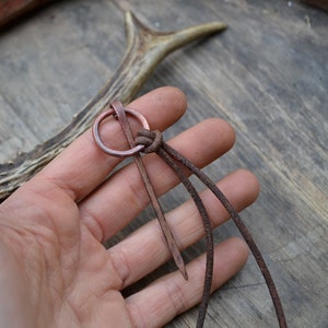 Pu&ograve; includere: Uno spillo in rame con un anello circolare nella parte superiore, infilato in un cordoncino di cuoio marrone. Lo spillo &egrave; tenuto in una mano su uno sfondo di legno.
