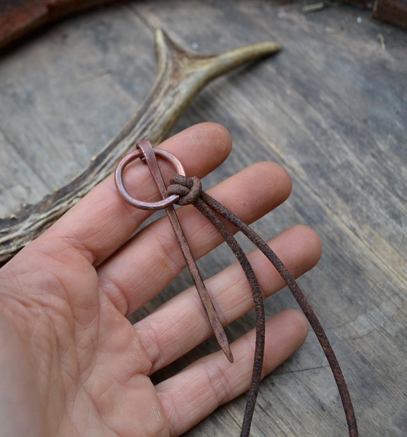 Pu&ograve; includere: Uno spillo in rame con un anello circolare nella parte superiore, infilato in un cordoncino di cuoio marrone. Lo spillo &egrave; tenuto in una mano su uno sfondo di legno.