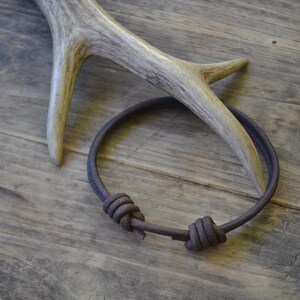 May include: A brown leather bracelet with two knots on a wooden surface. The bracelet is made of a thick, durable leather and has a simple, rustic design.