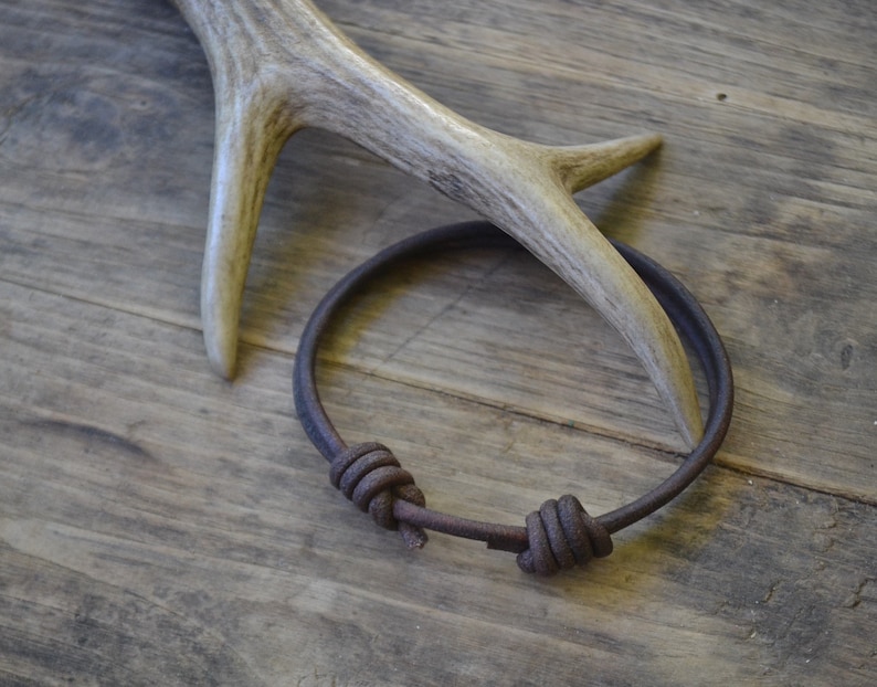 May include: A brown leather bracelet with two knots on a wooden surface. The bracelet is made of a thick, durable leather and has a simple, rustic design.