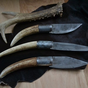 Könnte beinhalten: Drei handgefertigte Messer mit Hirschhorn-Griffen und Stahlklingen werden auf einer dunklen Oberfläche präsentiert. Die Klingen haben ein rustikales, abgenutztes Aussehen. Ein Geweih liegt über den Messern.