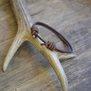 May include: A brown leather bracelet with a knotted design. The bracelet is displayed on a wooden surface.