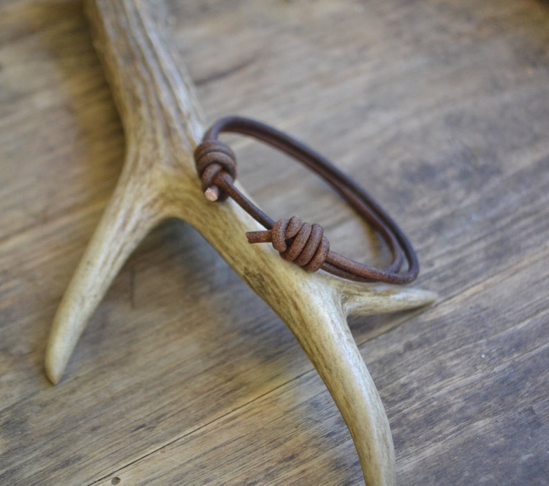 May include: A brown leather bracelet with a knotted design. The bracelet is displayed on a wooden surface.
