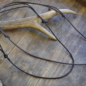 Può includere: Due cordini di cuoio, uno nero e uno marrone, sono legati insieme in un nodo. I cordini sono su una superficie di legno con un pezzo di corno di cervo.