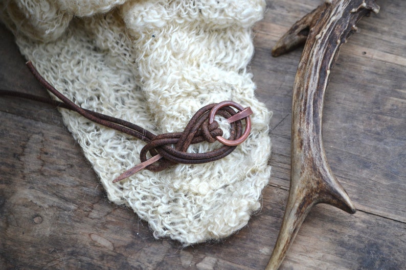 Pu&ograve; includere: Un cordoncino di cuoio marrone con un anello di rame e un'estremit&agrave; appuntita &egrave; legato in un nodo. Il cordoncino &egrave; avvolto attorno a un tessuto bianco e soffice e un corno di cervo marrone.
