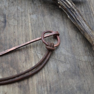 Pu&ograve; includere: Una collana con un ciondolo in rame composto da un lungo pezzo di metallo sottile e appuntito attaccato a un anello. La collana &egrave; realizzata con un cordoncino in pelle marrone.