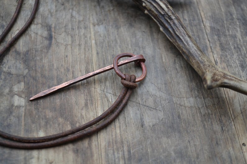 Pu&ograve; includere: Una collana con un ciondolo in rame composto da un lungo pezzo di metallo sottile e appuntito attaccato a un anello. La collana &egrave; realizzata con un cordoncino in pelle marrone.