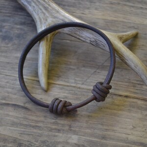 May include: A brown leather bracelet with two knotted accents. The bracelet is lying on a wooden surface with a piece of antler behind it.