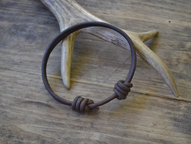 May include: A brown leather bracelet with two knotted accents. The bracelet is lying on a wooden surface with a piece of antler behind it.