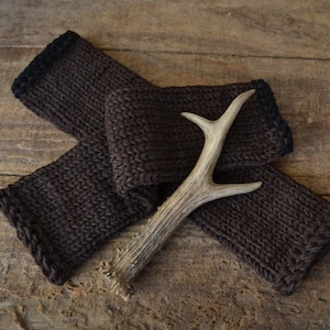 May include: A pair of brown knitted arm warmers with black trim, folded and arranged with a light brown antler on a wooden surface.