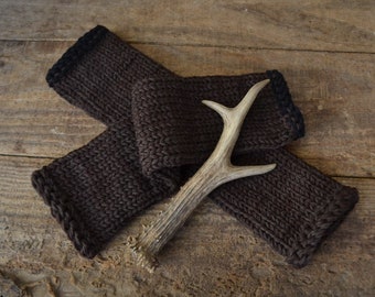 May include: A pair of brown knitted arm warmers with black trim, folded and arranged with a light brown antler on a wooden surface.