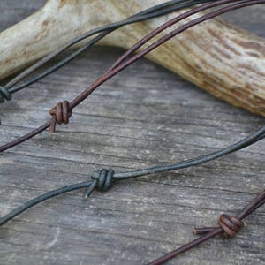 Peut inclure: Deux cordons en cuir noués, l'un brun et l'autre noir, sont présentés sur une surface en bois. Les cordons sont disposés en croisillon, le cordon brun au-dessus du cordon noir.