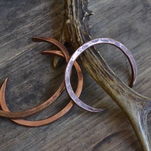 Pu&ograve; includere: Tre braccialetti in rame con texture martellate, impilati uno sopra l'altro, appoggiati su una superficie di legno con un corno di cervo.