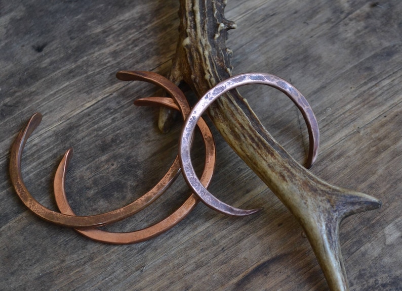 Pu&ograve; includere: Tre braccialetti in rame con texture martellate, impilati uno sopra l'altro, appoggiati su una superficie di legno con un corno di cervo.