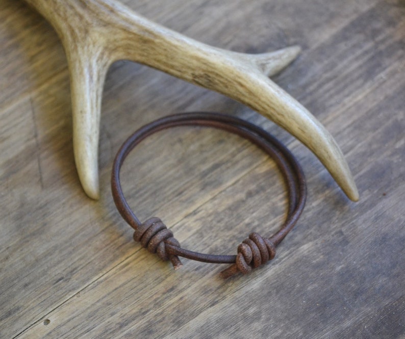 May include: A brown leather bracelet with two knots on a wooden surface.